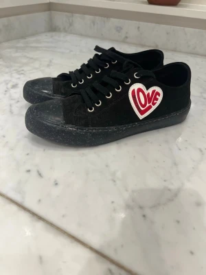 Svarta Love Moschino sneakers - Säljer ett par svarta sneakers från Love Moschino med klassisk låg siluett. Skorna har svart tyg, svarta snören och en vit och röd hjärtpatch med texten 'LOVE' på sidan. Snygga metallöljetter och gummisula ger en cool streetstil. Köptes i butik för ca 1 400kr. Använt endast  2 gånger.                         Storlek 40, lite små i storlekarna!