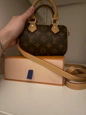 Louis Vuitton Mini Monogram Axelväska - Säljer en liten axelväska från Louis Vuitton i klassiskt brunt monogrammönster. Väskan har beige handtag och axelrem med guldfärgade detaljer. Perfekt för dig som gillar lyxiga accessoarer och vill ha något unikt och trendigt.