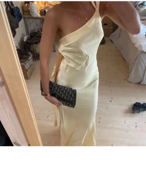 Ljusgul oneshoulder långklänning - Snygg ljusgul långklänning med oneshoulder-design och knyt i midjan. Klänningen har en elegant och stilren look, perfekt för dig som vill sticka ut. Materialet ser ut att vara mjukt och följsamt, vilket ger en fin passform.