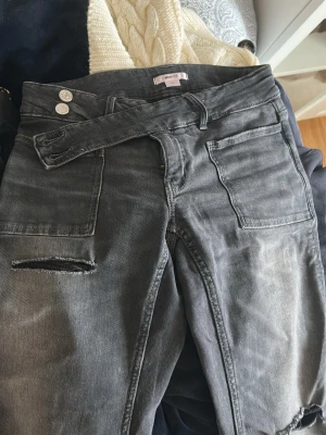 Svarta slitna jeans med stora fickor - Säljer ett par svarta jeans med tvättad look och slitningar på benen. Jeansen har stora fyrkantiga fickor fram och dubbla knappar i midjan. Raka ben och cool streetstil, perfekta för dig som gillar en avslappnad vibe.