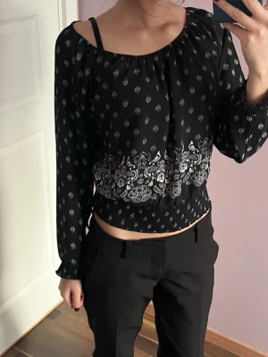 Svart off shoulder topp med mönster - Trendig svart off shoulder topp med vita småmönster och dekorativt blommigt band nedtill. Långärmad modell med resår vid ärmslut och midja. Kan bäras både på axlarna och off shoulder för olika looks. Luftigt material, perfekt till sommaren.