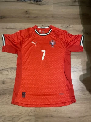 Portugal fotbollströja Puma röd - Säljer en röd Portugal fotbollströja från Puma med nummer 7 och Ronaldo på ryggen. Tröjan har vita och gröna detaljer vid kragen och ärmarna samt Portugal-emblem på bröstet. Tillverkad i lätt och ventilerande material, perfekt för träning eller match. Slim fit 