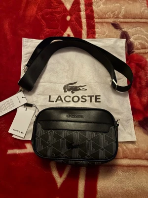 Svart axelväska från Lacoste - Snygg svart axelväska från Lacoste med justerbar axelrem. Perfekt för dig som vill ha en stilren och modern look.