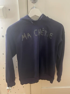 Mörkblå hoodie med text Ma Chérie - Mörkblå hoodie med huva och dragsko. På bröstet står det 'Ma Chérie' i glittriga stenar. Mjuk insida och ribbade muddar vid ärmslut och nederkant. Perfekt för en chill och trendig look.