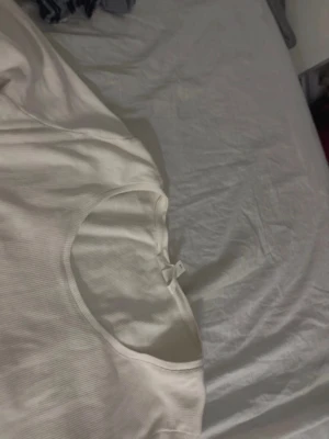 Vit ribbad långärmad topp - Säljer en enkel och stilren vit långärmad topp med rund halsringning. Toppen är ribbad och har en liten rosett vid halsen för en söt detalj. Perfekt att matcha med jeans eller kjol för en clean look.