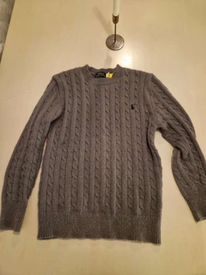 Grå kabelstickad tröja Polo Ralph Lauren - Kabelstickad grå tröja från Polo Ralph Lauren med rund halsringning och ribbade muddar. Tröjan har det klassiska broderade logotypen på bröstet och är tillverkad i ett mjukt, värmande material. Perfekt för lager-på-lager-stil.