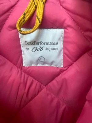 Rosa dunjacka från Peak Performance - Snygg rosa dunjacka från Peak Performance med quiltat mönster och dragkedja framtill. Jackan har en liten Peak Performance-logga på bröstet och är perfekt för kalla vinterdagar. Lätt och smidig att bära, med sportig känsla.