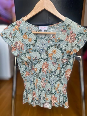Blommig topp från Vintage Stories - Supersöt blommig topp från Vintage Stories i ljusgrönt med volangkant och korta ärmar. Toppen har smock i midjan för en feminin siluett och ett romantiskt blommönster i orange, rosa och grönt. Perfekt för dig som gillar vintage vibes!