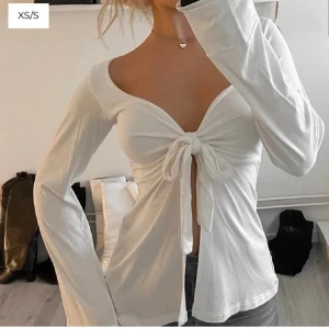 Vit omlottopp med knyt framtill - Trendig vit topp med omlottdesign och knyt framtill. Djup v-ringning och långa ärmar ger en snygg och feminin look. Perfekt att styla med jeans eller kjol för en cool outfit.