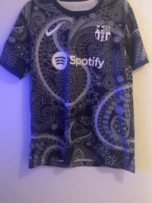 Barça x Spotify paisleymönstrad tröja - Snygg FC Barcelona-tröja från Nike med paisleymönster i svart och grått. Tryck med Spotify-logga på bröstet och klubbmärke. Korta ärmar och rund hals. Perfekt för dig som vill sticka ut på träningen eller matchen.