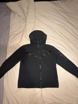 Svart Nike Tech Fleece hoodie - Säljer en svart Nike Tech Fleece hoodie med dragkedja och huva. Hoodien har snygga detaljer med svarta band över bröstet och en ficka med dragkedja på ärmen. Perfekt för en sportig och stilren look.