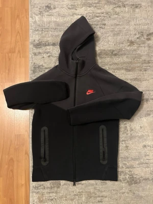 Svart Nike hoodie med dragkedja - Svart hoodie från Nike med hel dragkedja och huva. Har två svarta dragkedjeförsedda fickor framtill och en röd Nike-logga på bröstet. Tillverkad i mjukt material som känns skönt mot huden. Perfekt för en sportig och avslappnad stil.
