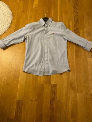 Ljusblå skjorta från Morris - Klassisk ljusblå skjorta från Morris med button down-krage och vita knappar. Skjortan har en liten röd broderad detalj på bröstet och är tillverkad i bomull.  Tvecka inte på att skriva 