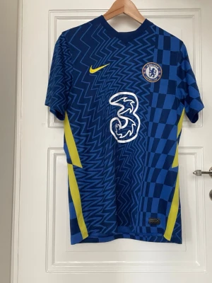 Chelsea FC matchtröja blå Nike - Chelsea FC matchtröja i blått med grafiskt mönster och gula detaljer. Tröjan har klubbmärke på bröstet, Nike-logga och stor vit sponsorlogga framtill. Tillverkad i lätt och ventilerande material, perfekt för fotboll eller träning.