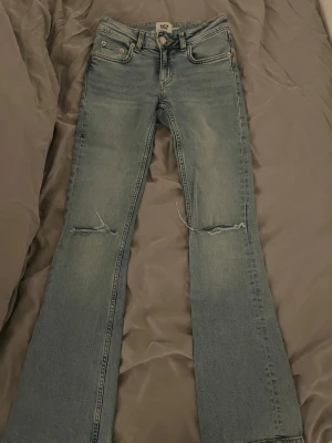 Bootcut jeans med slitningar från 157 - Säljer ett par ljusblå bootcut jeans från 157 med slitningar över knäna. Jeansen har klassisk femficksmodell, låg midja och är tillverkade i denim. Perfekta för en avslappnad och trendig look.