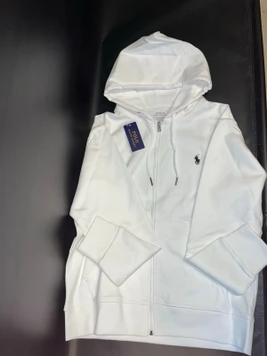 Vit hoodie från Polo Ralph Lauren - Säljer en fräsch vit hoodie från Polo Ralph Lauren med dragkedja framtill och klassisk huva. Tröjan har den ikoniska lilla loggan broderad på bröstet och är tillverkad i mjukt bomullsmaterial. Perfekt för en clean och stilren look.