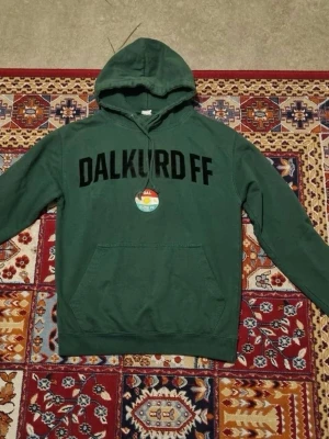 Grön Dalkurd FF hoodie - Mörkgrön hoodie med stor svart DALKURD FF-text över bröstet och klassisk känguruficka framtill. Huva med dragsko och ribbade muddar. Perfekt för dig som vill representera ditt favoritlag på ett avslappnat sätt.