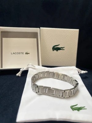 Silverfärgat armband från Lacoste - Stilrent armband i från Lacoste med rektangulära länkar och diskreta mönsterdetaljer. Levereras i original ask och tygpåse med klassiska Lacoste-loggan. Perfekt accessoar för dig som gillar en clean och modern look. Rostfritt och repfrit, ca 20cm justerbart. Nyskick