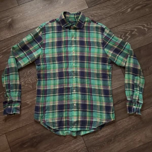 Ralph Lauren linneskjorta - Klassisk långärmad linneskjorta i grön, blå och vit rutigt mönster från Polo Ralph Lauren. Skjortan har knappar hela vägen, krage och rundad nederkant. Tillverkad i mjuk bomull som känns skön mot huden. Perfekt för en avslappnad men ändå stilren look. 100% linne, lite nopprig därav priset annars bra skick