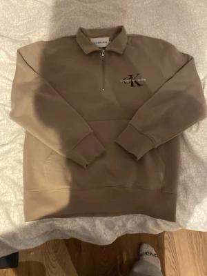 Beige sweatshirt Calvin Klein Jeans - Snygg beige sweatshirt från Calvin Klein Jeans med halv dragkedja och krage. Tröjan har ribbade muddar vid ärmslut och nederkant samt en stor CK-logga på bröstet. Perfekt för en avslappnad och trendig look.