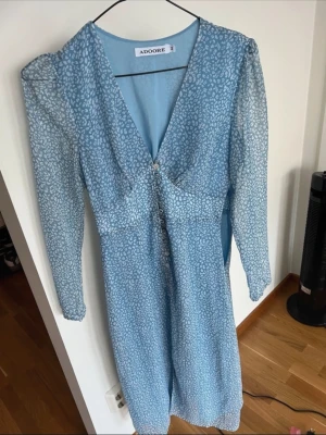 Blå mönstrad klänning från Adoore - Supersöt blå klänning från Adoore med v-ringning, långa ärmar och knappar framtill. Klänningen har ett ljust, småmönstrat tryck och en feminin passform som markerar midjan. Perfekt för dig som gillar stilrena men ändå lekfulla plagg.