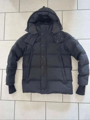 Canada goose wyndham - Snygg svart pufferjacka med stor huva och breda kanaler. Jackan har ribbade muddar vid ärmsluten och tryckknappar framtill. Märke syns på ärmen och jackan har en modern, boxig passform. Perfekt för kalla dagar och riktigt trendig look.