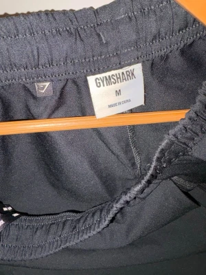 Svarta träningsshorts från Gymshark - Säljer ett par svarta träningsshorts från Gymshark i storlek M. Shortsen har elastisk midja och är tillverkade i ett lätt och snabbtorkande material, perfekta för gymmet eller löprundan. Diskret logga på benet och enkel, sportig design.