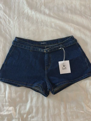 Mörkblå jeansshorts från Gina Tricot - Mörkblå denimshorts från Gina Tricot med hög midja och bälte med silverdetaljer. Klassisk femficksmodell och snygga sömmar. Perfekta för sommardagar och enkla att matcha med olika toppar.