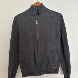 Mörkgrå ziptröja från Esprit - Mörkgrå stickad ziptröja från Esprit med hög krage och ribbade muddar. Tröjan har två fickor framtill och en diskret Esprit-logga vid nederkanten. Perfekt för lager på lager och enkel att matcha med olika outfits. Längd: 62cm, P2P: 42cm