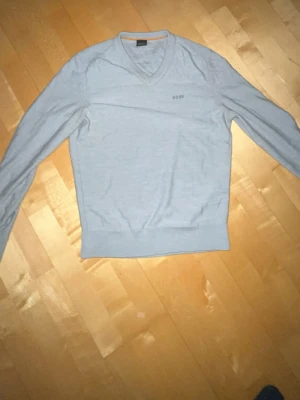 Grå v-ringad ull tröja från BOSS - Snygg grå v-ringad ull tröja från BOSS med diskret broderad logga på bröstet. Tröjan är stickad i mjuk ull och har ribbade muddar vid hals, ärmslut och nederkant. Perfekt för dig som gillar stilren och klassisk look..