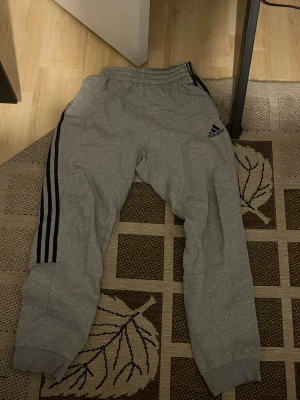 Gråa Adidas mjukisbyxor med svarta ränder - Gråa mjukisbyxor från Adidas med klassiska svarta ränder längs sidorna och Adidas-logga på vänster lår. Byxorna har resår i midjan och vid bensluten för en skön passform. Perfekta för chill eller träning. Storlek s