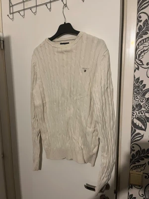 Vit kabelstickad tröja från Gant - Kabelstickad vit tröja från Gant med rund halsringning och diskret logga broderad på bröstet. Tröjan har ribbade muddar vid ärmslut och nederkant, och är tillverkad i mjukt stickat material. Perfekt för en klassisk och stilren look. Passar storlek M 