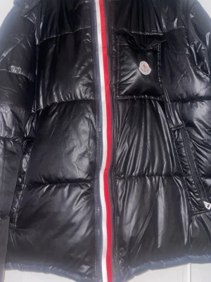 Svart Moncler dunjacka med röd/vit dragkedja - Snygg svart puffer dunjacka från Moncler med glansig finish. Jackan har en röd och vit rand längs dragkedjan, bröstficka med Moncler-logga och praktiska sidofickor. Perfekt för kalla dagar och ger en riktigt clean look.