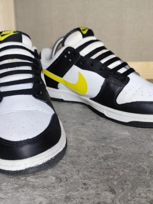Nike Dunk Low svart/vit/gul - Nike Dunk Low sneakers i svart och vitt läder med en iögonfallande gul Swoosh på sidan. Klassisk låg siluett med svart yttersula och vita detaljer. Skorna har svarta snören och vit plös med gul Nike-logga. Perfekt för dig som gillar streetwear och vill sticka ut.