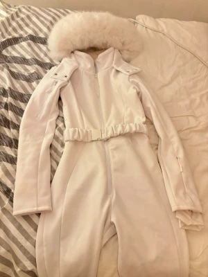 Vit overall med fuskpäls och bälte - Vit overall med dragkedja framtill, fluffig fuskpäls vid kragen och elastiskt bälte i midjan. Långa ärmar med dragkedjeficka och detaljerade axelpartier. Använd endast en gång, köpt från asos i storlek S, priset går att diskuteras