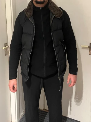 Svart dunväst från Parajumpers - Snygg svart dunväst från Parajumpers med dragkedja och två fickor framtill. Kragen är fodrad med mörkbrun päls. Perfekt att slänga över en hoodie eller tröja när det är kyligt ute. Storlek M Finns kvitto