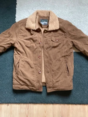 Brun teddyjacka med manchester - Säljer en brun teddyjacka från Rocky Mountain med mjukt teddyfoder och bred krage. Yttermaterialet är manchester och jackan har knappar framtill, bröstficka med lock samt två sidofickor. Perfekt för dig som gillar retrostil och vill ha något riktigt mysigt.