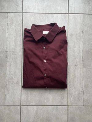 Mörkröd skjorta, tiger of Sweden - Stilren mörkröd skjorta från Tiger of Sweden med klassisk krage och knappar framtill. Skjortan är långärmad och tillverkad i mjuk bomull, perfekt för en clean och modern look. Vid frågor eller funderingar skriv gärna! 