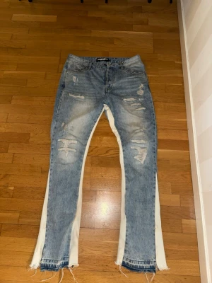Blå slitna jeans med vita paneler - Unika blå jeans med slitningar och råa kanter, samt vita tygpaneler längs benen. Jeansen har en rak passform och klassisk femficksdesign. Perfekta för dig som vill sticka ut med en edgy streetstil.