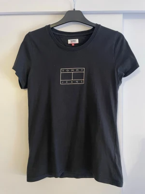 Svart t-shirt från Tommy Jeans - Svart t-shirt från Tommy Jeans med klassisk rund hals och korta ärmar. Framtill finns en stilren logga i guld.