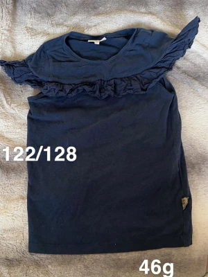 Marinblå topp med volangdetalj - Snygg marinblå topp med korta ärmar och volangdetalj över bröstet och axlarna. Enkel rund halsringning och rak passform. Perfekt för dig som gillar stilrena plagg med en twist.🌸 Fint använt skick. Storlek 122/128