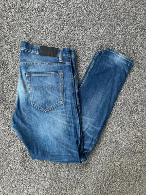 Blå tiger of sweden jeans - Snygga blå slim fit jeans med lätt slitna detaljer och klassisk femficksdesign. Jeansen har en mörkare tvätt med ljusare partier på lår och knän samt en svart patch bak i midjan. Perfekta för en avslappnad och trendig look.