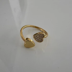 Guldfärgad ring med hjärtan - Trendig guldfärgad ring med öppen design och två hjärtan i ändarna. Ena hjärtat är slätt medan det andra är dekorerat med små gnistrande stenar för extra bling. Perfekt accessoar för att lyfta din stil med en söt och modern touch.