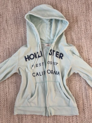 Hollister zip hoodie - Jättefin hoodie från Hollister köpt på sellpy. Den ser lite vit ut på bilden är men ljus turkos i verkligheten🩷storlek M men passar mig som är 165, Skriv om ni har några frågor osv💓