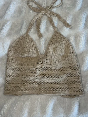 Beige virkad topp - Säljer en beige virkad topp med halterneck och snygg snörning i ryggen. Toppen är ärmlös och har ett bohemiskt, handgjort utseende som passar perfekt till sommaren. Supersnygg att styla med jeans eller kjol!