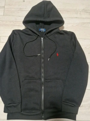 Svart hoodie från Polo Ralph Lauren - Klassisk svart hoodie från Polo Ralph Lauren med dragkedja, vit dragsko och den ikoniska röda loggan broderad på bröstet. Hoodien har kängurufickor och ribbade muddar. Tillverkad i mjuk bomullsblandning för skön komfort.