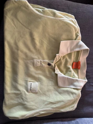 Ljusgul pikétröja från Hugo Boss - Snygg ljusgul pikétröja från Hugo Boss med vit krage och knappslå framtill. Tröjan har korta ärmar och en liten patch med logga på bröstet. Perfekt för en clean och fräsch stil.