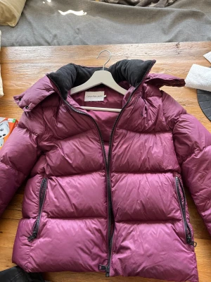 Lila pufferjacka Calvin Klein Jeans - Säljer en snygg lila pufferjacka från Calvin Klein Jeans i storlek M. Jackan har hög krage, huva och två fickor med dragkedja. Materialet är polyester och jackan har en quiltad look med breda paneler. Perfekt för kalla dagar och riktigt trendig färg.