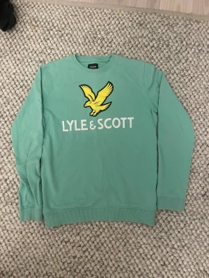 Mintgrön Lyle & Scott sweatshirt - Fräsch mintgrön sweatshirt från Lyle & Scott Junior med stor gul fågel och vit logga på bröstet. Tröjan har rund hals, ribbade muddar och är långärmad. Perfekt för dig som gillar stilrena och sportiga vibes.