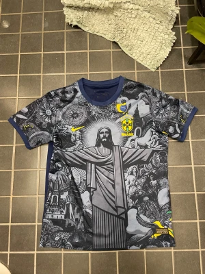 Nike Brasil Dri-Fit ADV t-shirt - Snygg Brasil t-shirt från Nike med grymt detaljerat grafiskt tryck på framsidan, bland annat Kristusstatyn och coola mönster. T-shirten är mörkblå med inslag av grått och gult, har korta ärmar och är gjord i lätt polyester med Dri-Fit ADV-teknologi. Perfekt för fotboll eller häng med kompisar.